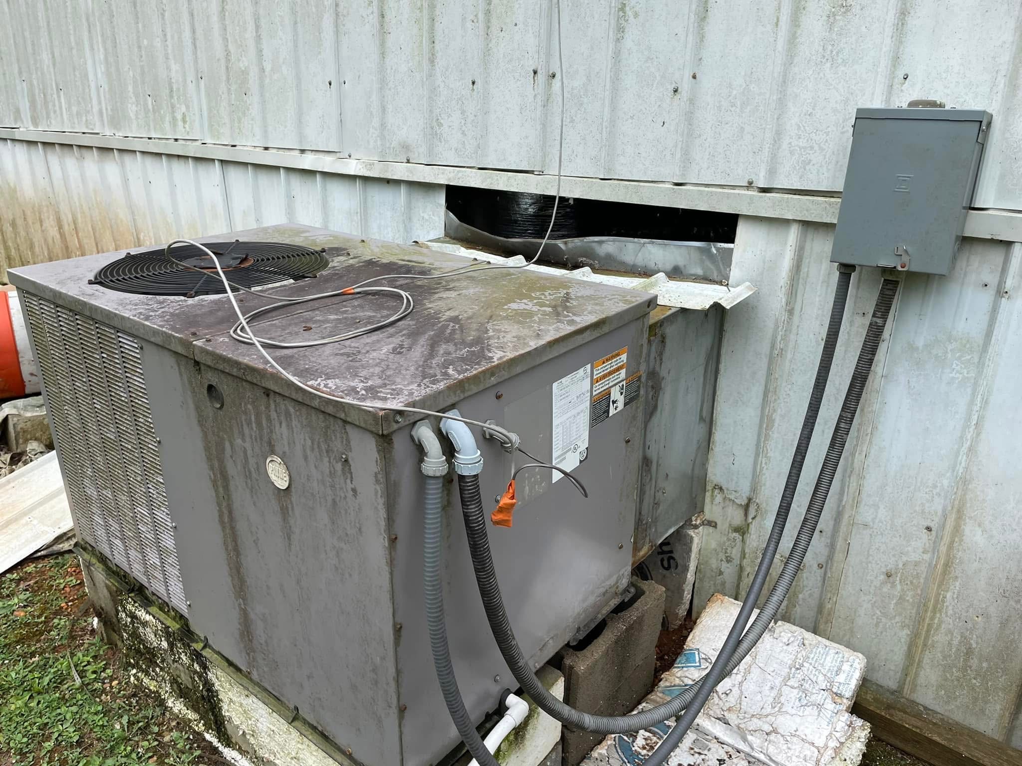 Worn, stained package unit with loose wiring and damaged ductwork beside a mobile home